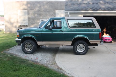 1996 Ford Bronco - A green 1996 Ford Bronco is parked in front of a garage, with a pink toy car visible inside.