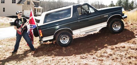 1993 Ford Bronco - A 1993 Ford Bronco is shown on a dirt hill, with a person holding a guitar and a flag nearby.
