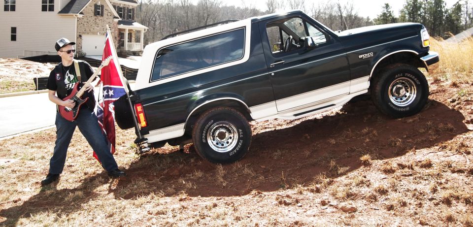 1993 Ford Bronco parked on a dirt incline with a person holding a guitar and a Confederate flag