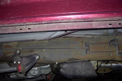 1994 Ford Bronco - Close-up of the rusty frame and suspension parts of a vehicle, with a red body panel overhead.