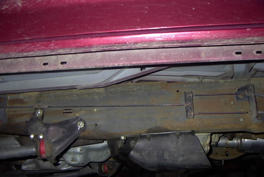 Underside view of a vehicle's frame and suspension components. Red body panel visible above