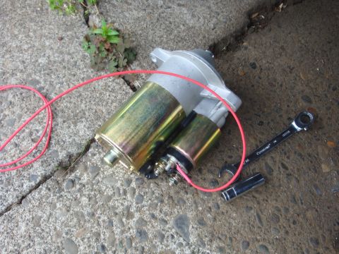 1989 Ford Bronco - A starter motor, red wiring, a wrench, and a socket are laid out on a cracked concrete surface.