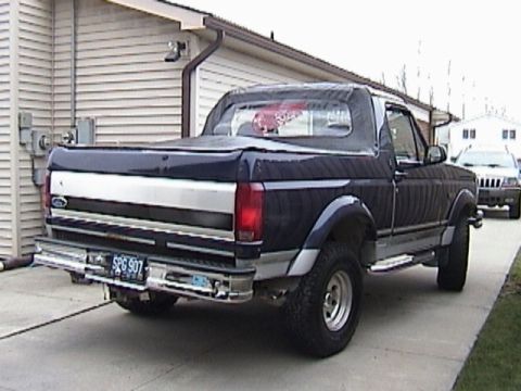 1995 Ford Bronco - The rear of a dark blue Ford Bronco is visible, parked in a driveway next to a house. A second vehicle is parked behind it.