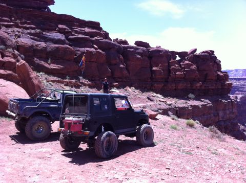 1987 Suzuki Samurai - Two modified off-road vehicles are parked on a dusty trail next to a large red rock formation.