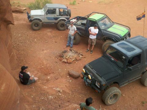 1987 Suzuki Samurai - Three off-road vehicles are parked on a dirt landscape, with people gathered around a campfire.