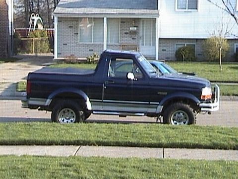 1995 Ford Bronco - A dark blue 1995 Ford Bronco is parked on the side of a street in front of a house, with another vehicle partially visible behind it.