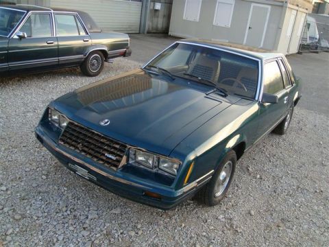 1991 Ford LTD Crown Victoria - A 1980s Ford Mustang coupe is parked on a gravel lot next to another sedan.