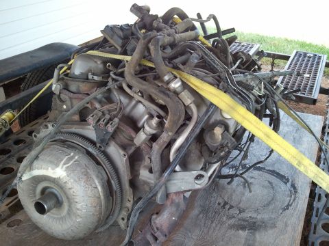 1989 Ford Bronco - A complex engine and transmission assembly is shown, secured by yellow straps on a wooden platform.