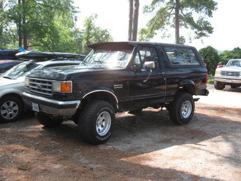 1994 Ford Bronco - A lifted black Ford Bronco is parked on a dirt lot, with trees and another vehicle in the background.