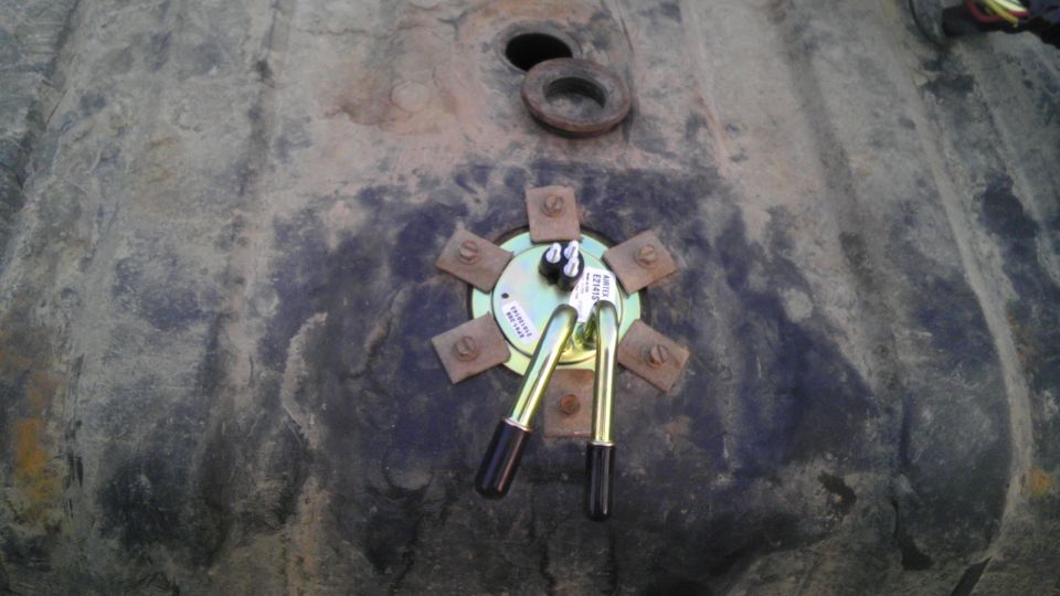 Close-up of a fuel pump assembly with two brass float arms attached to a metal plate, bolted onto a dark, textured surface that appears to be a fuel tank