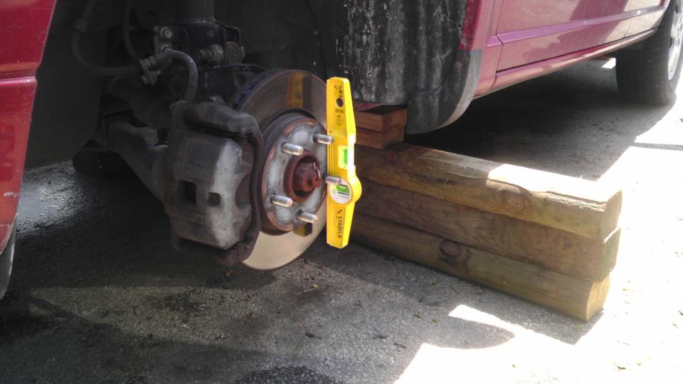 Car's front wheel assembly is raised on wooden blocks, with a yellow level tool attached to the brake rotor