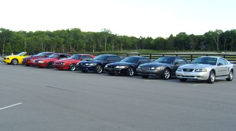 2007 Ford Mustang - Multiple generations of Ford Mustangs are lined up in a parking lot with a wooded background.