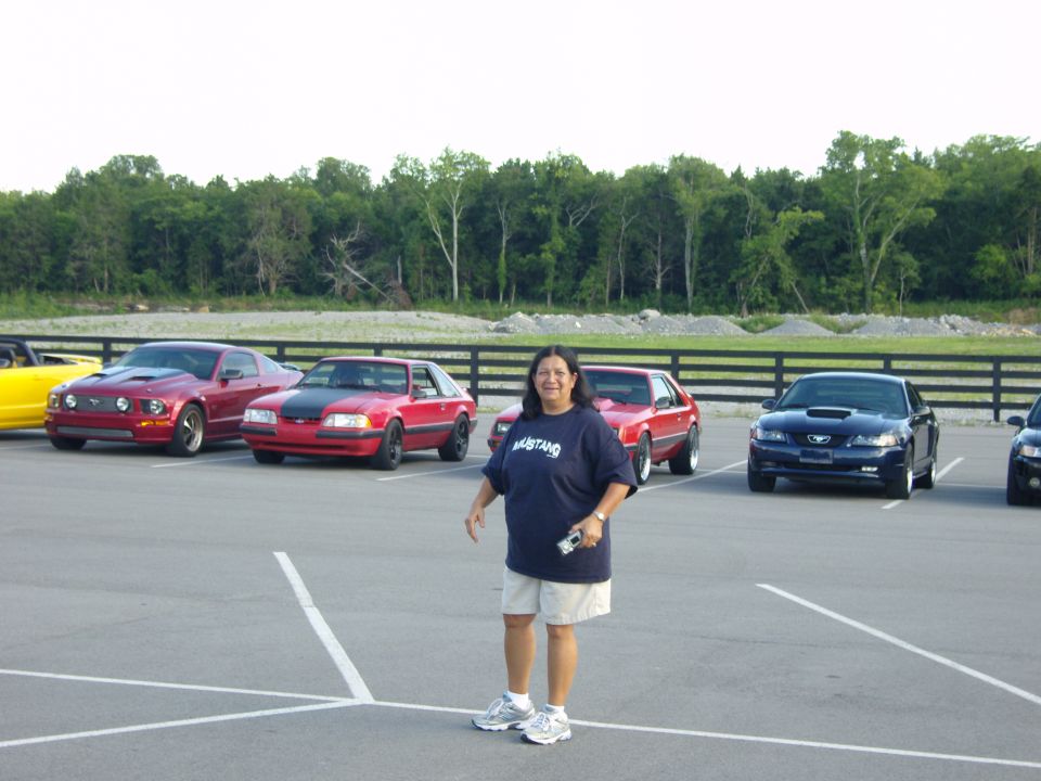 Woman in 'Mustang' shirt stands in parking lot with red, blue, and yellow Ford Mustangs