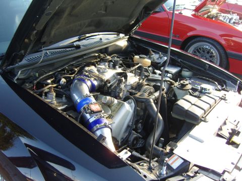 2007 Ford Mustang - The engine bay of a black car is shown with an aftermarket intake system. Another vehicle is parked behind it.