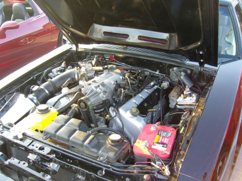 2007 Ford Mustang - The engine bay of a black car, modified with a supercharger, is shown with another red car in the background.