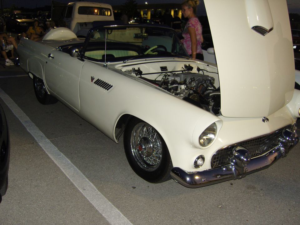 White 1955 Ford Thunderbird convertible with hood open, wire wheels, and a woman standing nearby