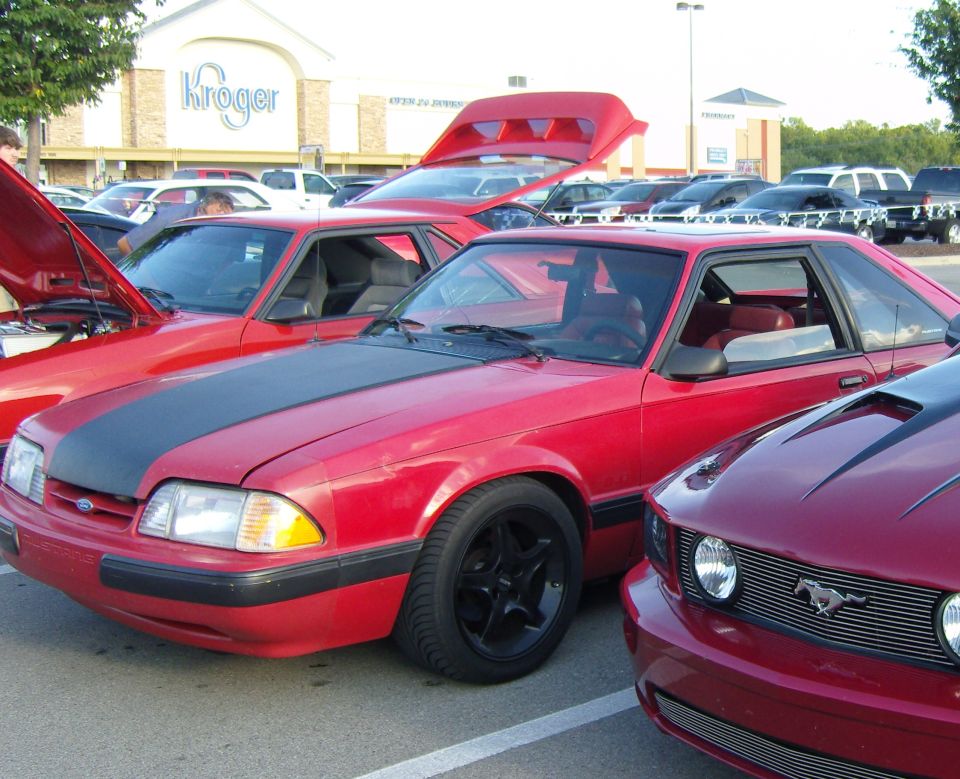 Red Ford Mustang with black hood stripe and black wheels, parked next to other cars
