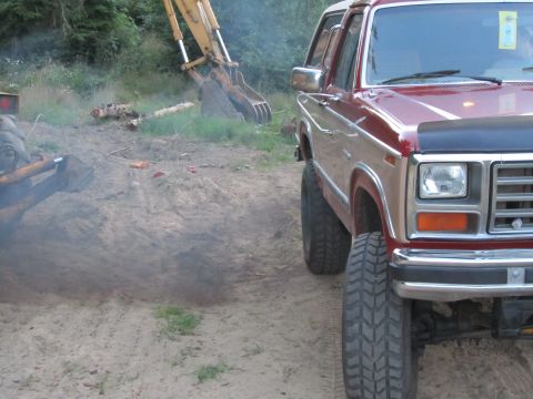 1984 Ford Bronco - A red and tan Ford Bronco is parked on a dirt lot with heavy machinery in the background.