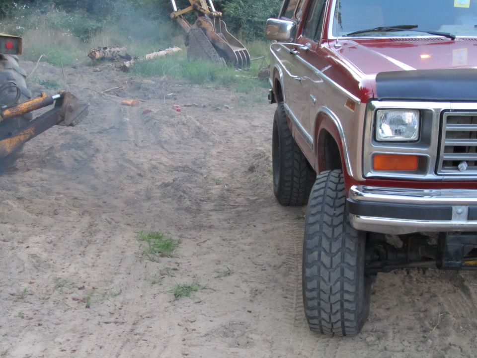 Red Ford Bronco with lifted suspension and large tires parked on dirt. Excavator in background