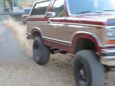 1984 Ford Bronco - A two-tone Ford Bronco is shown driving off-road, with a construction vehicle visible in the distance.