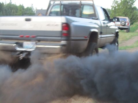 1984 Ford Bronco - A Dodge pickup truck is seen from the rear, spewing thick black exhaust smoke, with another vehicle visible in the distance.