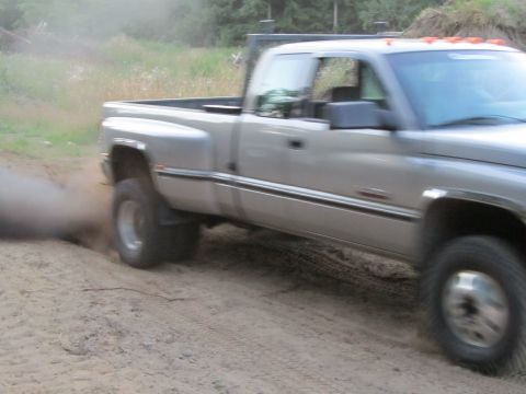 1984 Ford Bronco - A silver pickup truck with dual rear wheels drives on a dirt road, kicking up a cloud of dust.