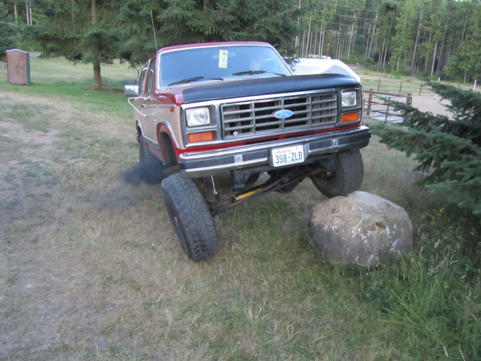 Red and tan Ford Bronco with lifted suspension, one tire on a large rock, emitting black smoke. License plate 398-ZLB