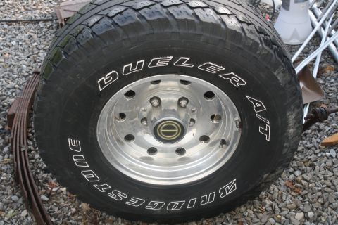 1996 Ford Bronco - A close-up of a Bridgestone Dueler A/T tire mounted on a chrome wheel, with a leaf spring visible to the side.