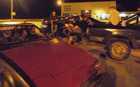 2006 Ford E350 - Several people are gathered around vehicles outside a service garage at night, with a tow truck in the background.