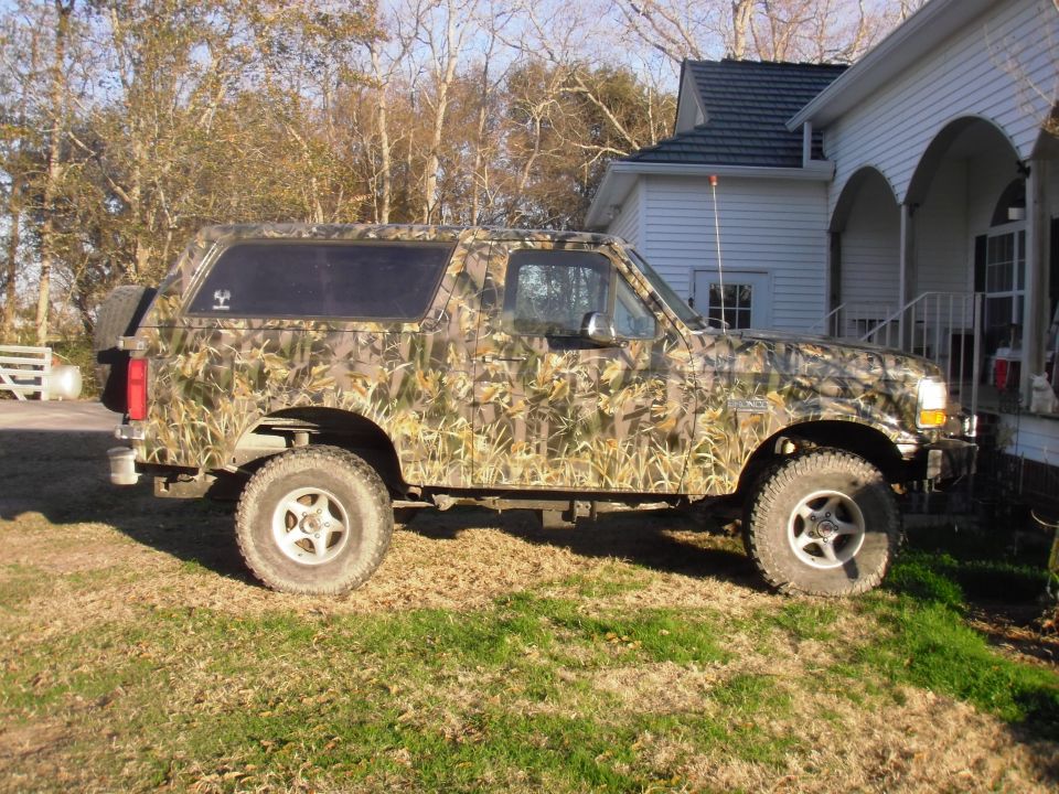 Camouflage-wrapped lifted Ford Bronco with big mud tires, side view, parked on grass next to a white house