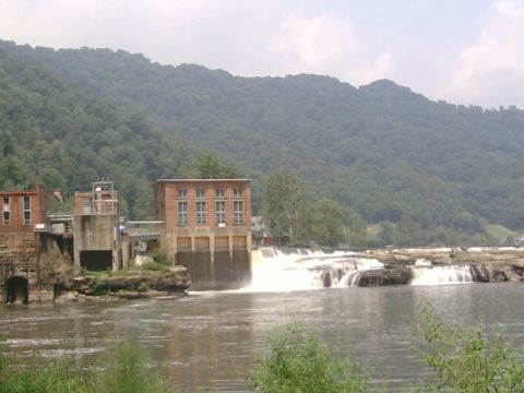 1985 Ford Crown Victoria LTD - A hydroelectric dam with a waterfall flows into a river, bordered by brick buildings and a lush, tree-covered mountain range.
