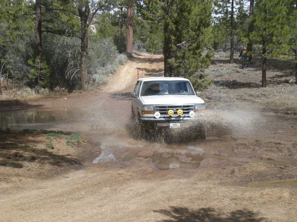 White Ford Bronco splashing through a muddy puddle on a dirt road, front view