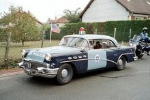 1991 Ford Crown Victoria - This vintage police car, likely from the 1950s, is seen driving down a street with a motorcycle following.