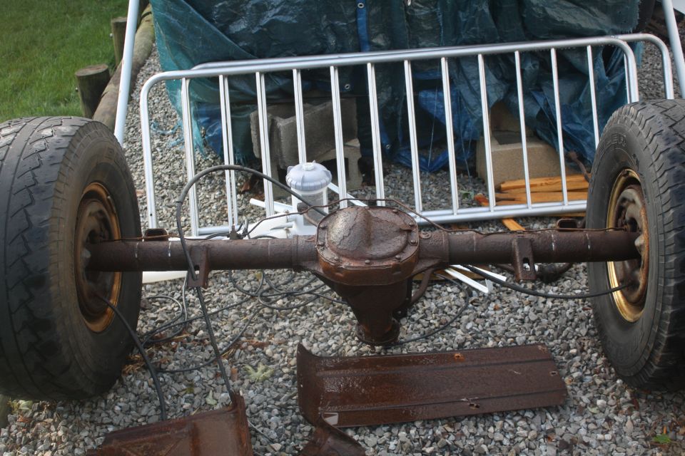 Rusty rear axle with two wheels, brake lines, and some metal trim pieces on gravel
