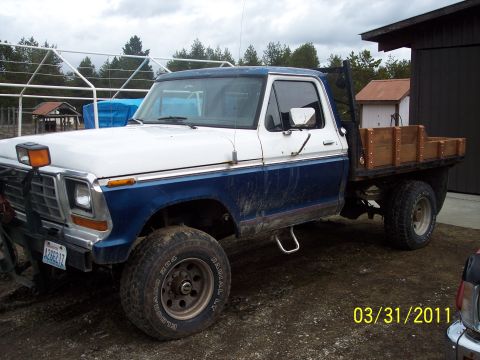 1973 Ford F-250 - Driver-side profile of a white-over-blue 4x4 Ford regular-cab pickup with a wood stake-side flatbed, black headache rack and a plow mount h…
