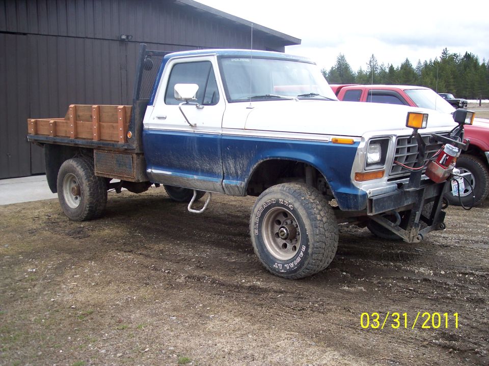 Blue and white 4x4 Ford flatbed pickup with plow mount, front 3/4 passenger side view