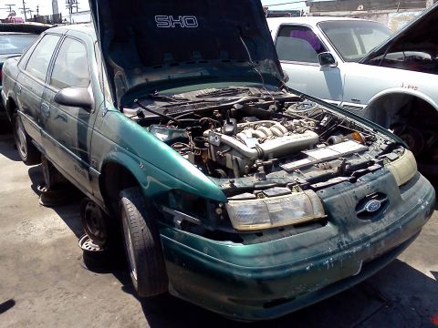 1993 Ford Taurus - A green Ford Taurus SHO sits in a junkyard with its hood open, revealing the engine compartment.
