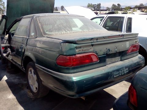 1993 Ford Taurus - Rear 3/4 view of a green 1993 Ford Taurus sedan in a junkyard, with the driver's door ajar.