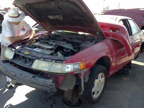1993 Ford Taurus - Man works on the engine of a red 1993 Ford Taurus SHO at a junkyard.