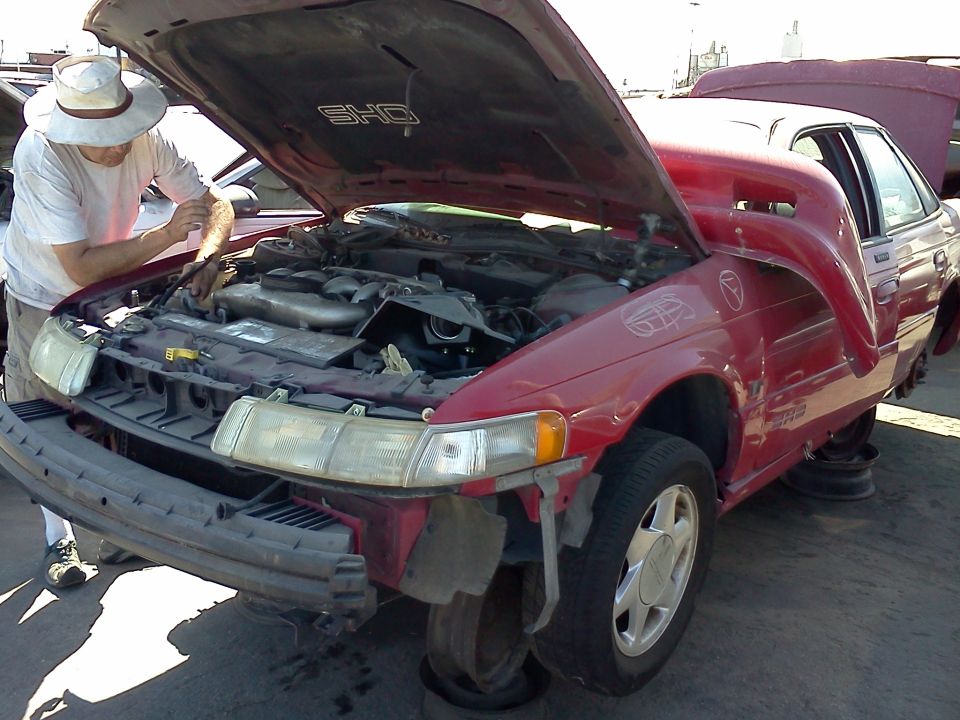 Man inspects engine of red 1993 Ford Taurus SHO with hood open. Front bumper is removed