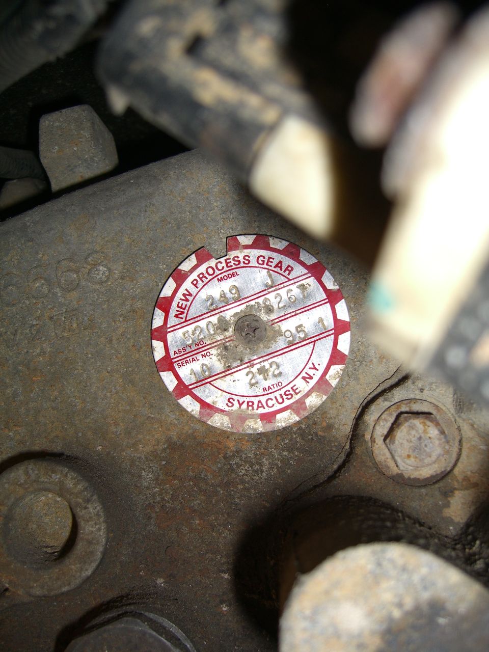 Close-up of a round red and white tag on a rusty metal part. Text reads 'NEW PROCESS GEAR', 'MODEL 249', 'SERIAL NO. 40', 'ASSY NO. 5204-95 1', 'RATIO 272', and 'SYRACUSE N.Y.'