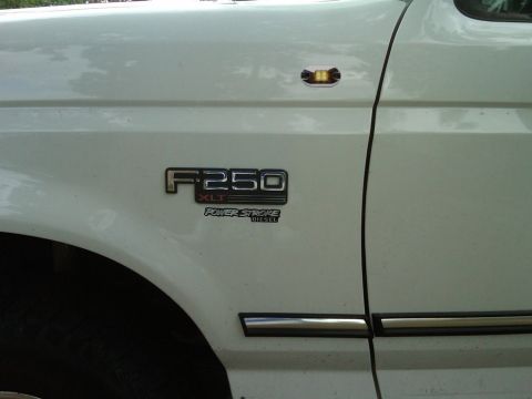 1994 Ford Bronco - Close-up of the front fender on a white Ford pickup showing the chrome F-250 XLT badge with a Power Stroke Diesel emblem beneath it. An amb…