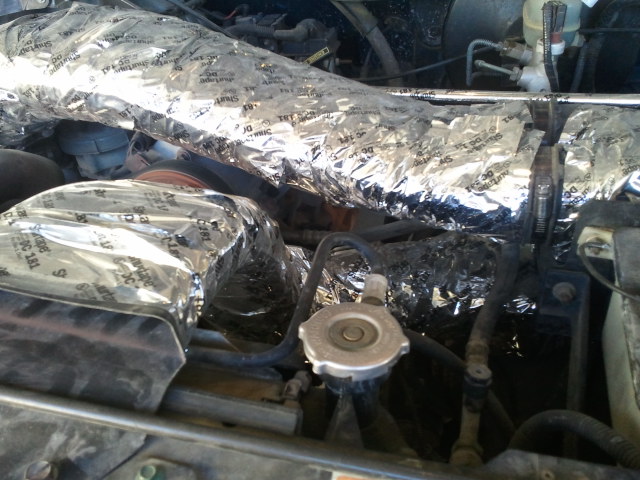 Engine bay with shiny insulation wrapped around hoses and parts. Radiator cap visible