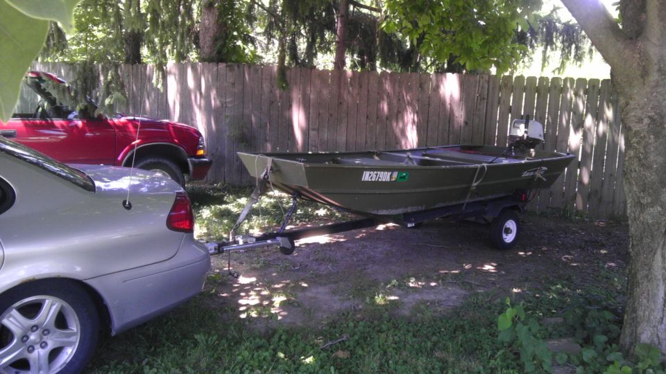 Green jon boat on trailer, hitched to a silver sedan, with a red truck behind it