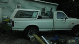 1989 Ford Bronco - A white pickup truck with a camper shell is parked outside a building.