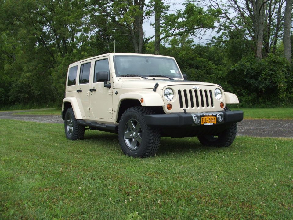 Tan four-door Jeep Wrangler parked on grass with trees in background. License plate reads FJE-4044