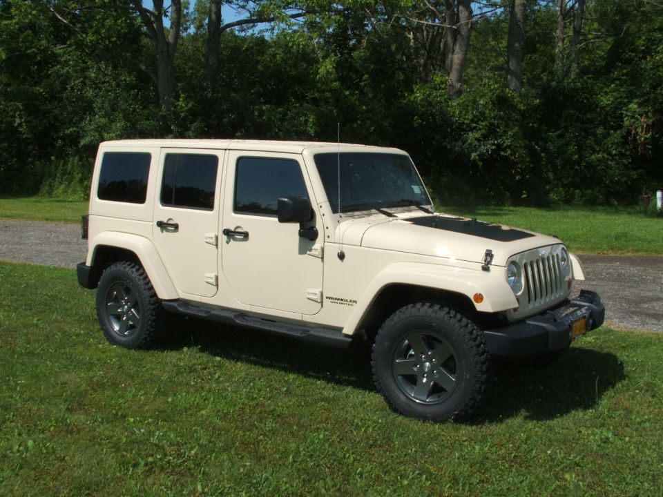 Tan 4-door Jeep Wrangler parked on grass with trees in background. Has black hood decal and dark wheels