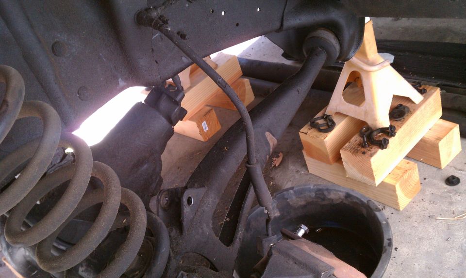 vehicle's front suspension supported by jack stands and wooden blocks, with a coil spring visible