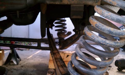 1990 Ford Bronco - The underside of a vehicle shows rusty suspension components, including coil springs and control arms, elevated on jack stands.