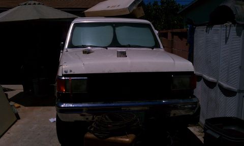 1990 Ford Bronco - A white Ford truck is parked in a driveway, partially covered by a patio umbrella and next to a storage unit.